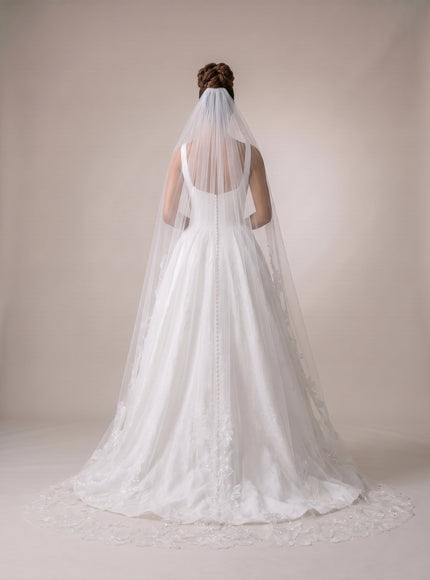 Person wearing a white wedding dress with a long veil against a plain background