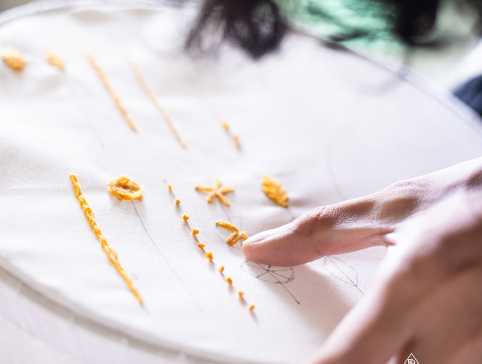 Close-up of hands working on embroidery with orange thread on a white fabric.